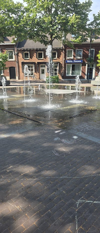 The image depicts a public square with a water fountain in operation, located in Horst, Netherlands. The square is paved with dark interlocking bricks, and there are chalk drawings on the ground, including circular patterns and lines. A row of brick buildings lines the square, featuring tiled roofs and multiple windows. One building has a blue sign that reads "HOTEL-PENSION". A bicycle is visible leaning against one of the buildings. Green trees with full foliage are situated behind the buildings, and the sky is partially visible. Sunlight casts shadows across the square.
