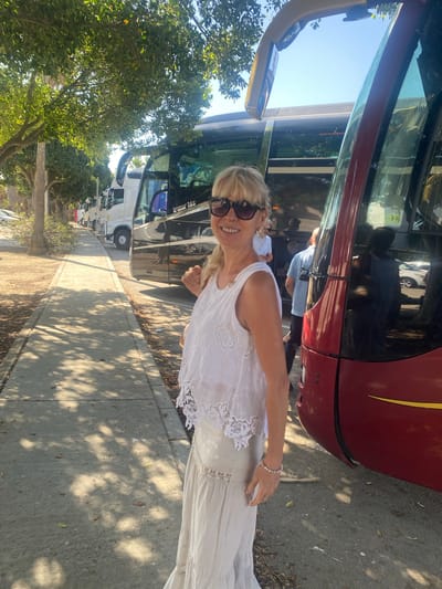 The image shows a woman in a white top and a long, light-colored skirt standing on a sidewalk next to a line of parked buses. She is wearing sunglasses and smiling. Behind her, a row of buses, including a white truck and several dark-colored passenger buses, are parked along the street. Trees with green foliage line the sidewalk, casting shadows on the ground. The scene appears to be outdoors in a sunny environment. The location is identified as Pol. Ind. La Palmosa, Spain.