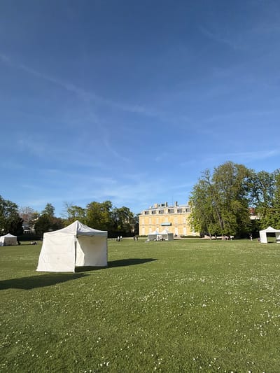 The image depicts a sunny scene in Rueil-Malmaison, France. A large lawn stretches out, dotted with small white flowers. A grand, yellow building stands in the background, partially obscured by trees. Several white tents are set up on the lawn. People are walking or standing near the building and the tents. The sky is clear and blue with faint clouds.