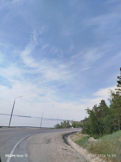 The image depicts a curved asphalt road bordering a body of water, possibly a river or lake, in Nizhny Bestyakh, Russia. The road is lined with streetlights and a guardrail. On the right side of the road, there is a slope covered with grass and trees, including pine and birch. The sky is blue with wispy white clouds. In the distance, along the water's edge, there are some industrial structures visible. A timestamp in the bottom right corner reads "07.07.2026 16:43", and a watermark in the bottom left corner reads "REDMI Note 13". There are no people visible in the image.