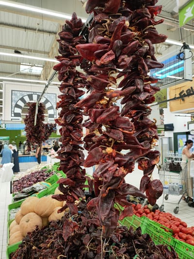 The image depicts a grocery store produce section with an abundance of dried chili peppers hanging from the ceiling. In the foreground, several strings of dark red, dried peppers are prominently displayed. Below them, green plastic crates are filled with various produce. One crate contains several large, light-colored pumpkins. Another crate holds a large quantity of red tomatoes. Further back, more green crates are visible, holding what appear to be red onions and other vegetables.

In the background, other shoppers are present. Two women, one in a blue hijab and a long blue garment, and another in a light-colored hijab and a dark dress, are standing near a produce display. Other individuals are seen browsing or pushing shopping carts. The store environment is characterized by bright overhead lighting, a tiled floor, and shelving units. A decorative archway with Islamic geometric patterns is visible on a wall in the mid-ground. The overall impression is a bustling supermarket produce aisle. The location is specified as Cebalat, Tunisia.