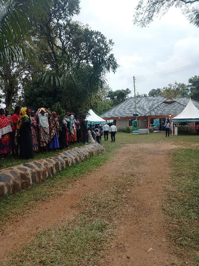 The scene depicts an outdoor gathering in Marenyo-Shianda ward, Kenya, under an overcast sky. In the foreground, a reddish-brown dirt path runs through sparse green grass, bordered on the left by a low stone wall approximately 0.5 meters high. To the left of this path, behind the stone wall, a line of approximately 20-30 individuals, predominantly women, are standing. They are dressed in various colorful patterned garments, with several wearing head coverings; some appear to be balancing items on their heads. Directly behind them are dense green trees and foliage.
In the midground, the dirt path extends towards a single-story building with a grey, multi-pitched tiled roof and light-colored walls. A utility pole with wires stands centrally in front of the building. Two white canopy tents are erected on the grassy area, one to the left and one to the right of the building. Several men, dressed in light shirts and dark trousers, are walking on the grassy area towards the building. Near the building, at least two banners are visible; one predominantly green, and another blue, with some text and images. The background consists of numerous large green trees, indicating a semi-rural or heavily vegetated environment surrounding the building. No direct interactions among the individuals are clearly visible from this perspective.