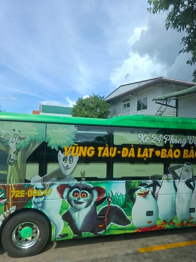 The image shows the side of a green bus with cartoon characters from the movie Madagascar painted on its exterior. The characters include a giraffe, a lemur, and several penguins. The bus has windows with blinds, and some text is visible on the side, including "Xe 24 Phong V" and "VUNG TAU-DA LAT BAO BAO". A license plate with "72E-00647" is also visible. The bus is parked on a concrete surface with a yellow line. In the background, there is a building with a white facade and a gray roof, and trees. The sky is blue with some white clouds. This scene is located in the city of Tân Phú 2, Vietnam.