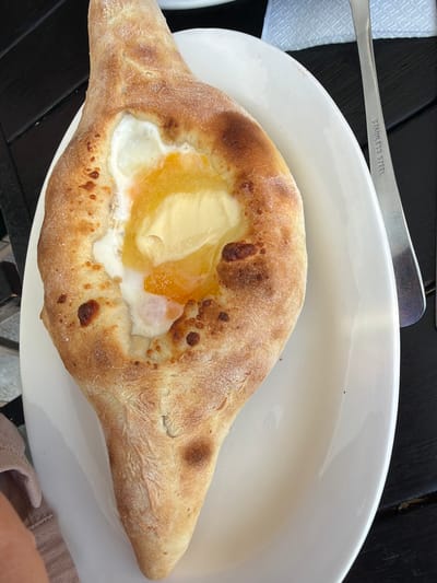 The image shows a Khachapuri, a Georgian cheese-filled bread, served on a white oval plate. The Khachapuri is boat-shaped and filled with melted cheese, a fried egg, and a pat of butter. The bread has a golden-brown crust with some darker, charred spots. A fork with "STAINLESS STEEL" engraved on it is partially visible on the right side of the plate. The plate rests on a dark wooden table. A crumpled white napkin is visible in the upper right corner of the image. A pinkish-beige napkin or cloth is on the left side of the plate, partially obscuring the table. The background is blurred, but it appears to be an outdoor setting, likely a cafe or restaurant, possibly in Tsandrypsh, Abkhazia, given the context. There are no people clearly visible in the image.