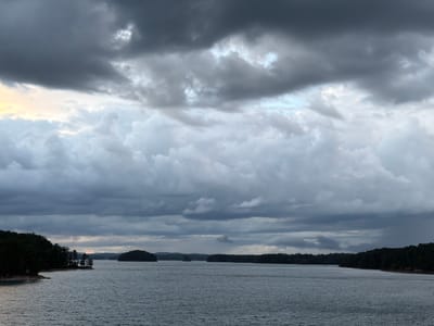 The image depicts a gray and overcast day over a large body of water, likely Lake Lanier in Sugar Hill, Georgia, United States. Dense forests line both sides of the water, forming a shoreline with occasional islands. The sky is filled with heavy, layered clouds, with some faint hints of light breaking through. The water is calm and reflects the gray sky. There are no visible people in the image.