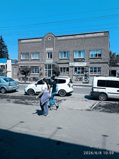 The image shows a street scene in Lebedyn, Ukraine, on June 8, 2026, at 10:59 AM. A two-story brick building with arched window details and decorative brickwork along the roofline stands on the right side of the frame. The building has multiple windows on both floors, some with white blinds or curtains. Several businesses appear to be located on the ground floor, indicated by signage, including "BLOOMBER" and other unreadable text in what appears to be Cyrillic. The building is situated on a paved street with a sidewalk in front.

Two women are walking on the sidewalk in the foreground, moving from left to right. The woman on the left is wearing a light purple top and light blue pants. The woman on the right is wearing a black t-shirt and blue jeans. Both are walking in the same direction, with the woman on the left slightly ahead.

Several vehicles are parked or driving on the street. A silver sedan is parked on the left side of the street. A white minivan is stopped in the street in the middle ground, and another white van is parked on the right side of the street. There are trees and power lines visible in the background. The sky is a clear blue. The overall impression is of a sunny day in a small urban or town setting.
