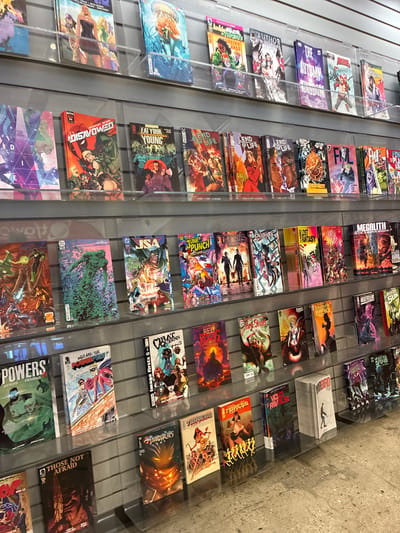 The image shows a close-up of several rows of comic books displayed on clear shelves in a store. The comic books are colorful and feature various illustrations and titles. Some of the visible titles include "The Disavowed," "Eat Your Young," "End of Life," "Junk Punch," "Lost Fantasy," "Red Skull," "Thunderbolts Lost," and "Venom Kill Spiders." The comic books are arranged in a neat, organized manner, with their covers facing forward. The background consists of a grey, textured wall, and the flooring appears to be concrete. There are no people or other subjects visible in the image. The lighting is bright and even, suggesting an indoor retail environment. The overall scene depicts a section of a comic book store.