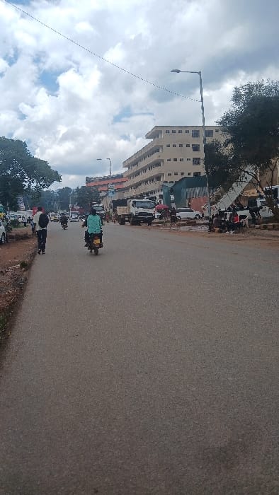 The image displays a daytime street view within Kisii Central ward, Kenya. The scene features a paved asphalt road stretching into the distance under a partly cloudy sky. A single individual walks away from the viewer on a dirt shoulder on the left side of the road, wearing a white shirt and dark trousers, carrying a dark backpack. A person on a motorcycle, wearing a light blue/green jacket, rides in the center of the road, also moving away from the viewer.
On the right side of the road, a multi-story, light-colored building with numerous rectangular windows rises, appearing to be at least five floors high. Further along the right, a white dump truck is visible, facing away, followed by several other vehicles, including white cars and a yellow mini-bus (matatu). Multiple dark street light poles line the right side. In the mid-ground on the right, several individuals are observed sitting and standing near damaged or partially constructed structures featuring exposed corrugated metal, with additional white vehicles parked in this area. Trees and shrubs are present on both sides of the road.