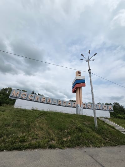 A large, angled sign with bold, light-brown Cyrillic lettering spelling "МОСКВА-ВЛАДИВОСТОК" (Moscow-Vladivostok) stands prominently on a grassy hill. The sign is supported by a white, angled structure, with the letters themselves appearing to be individual blocks. To the right of the sign, a tall, weathered lamppost with multiple light fixtures extends upwards, with a single black wire running across the sky. Behind the sign and lamppost, a structure resembling a stylized flag or monument with horizontal red, blue, and white stripes is visible, attached to a beige pillar. The background consists of a dense line of green trees and an overcast, cloudy sky. To the right of the sign, a concrete staircase leads up the hill. The foreground shows a gray, asphalt road. The scene is likely a city limit or a significant route marker in Nikolaevka, Russia.
