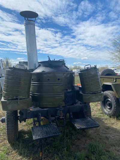 The image depicts a military field kitchen set up outdoors, likely on a grassy field near the city of Kumakskoe, Russia. The field kitchen consists of a large, dark green, trailer-mounted structure with several cylindrical containers, potentially cooking vessels, visible on top. A long, silver chimney with a circular cap extends upwards, suggesting it is in use. The structure has what appears to be fold-down metal platforms on the sides. A military vehicle is partially visible in the background. The sky is filled with white, puffy clouds and there is a blue sky behind them.