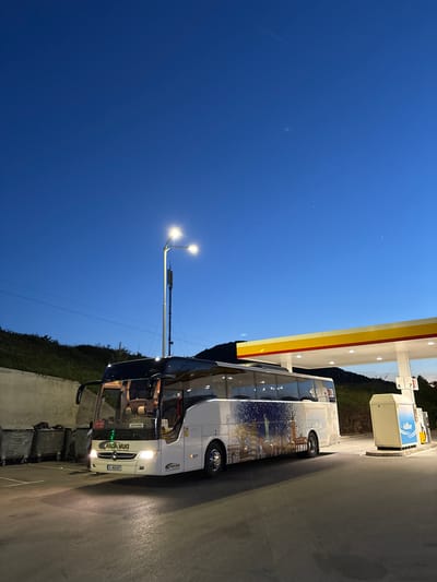 A white tour bus is parked at a Shell gas station in Kalotina, Bulgaria, likely at night given the dark blue sky. The bus has a large image printed on its side depicting landmarks like the Statue of Liberty, likely representing New York City. The license plate on the bus reads "K 4624 BT". A streetlight illuminates the front of the bus, and several trash bins are beside the bus. The Shell gas station is visible with its yellow and red canopy.