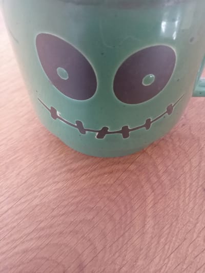 A close-up, eye-level shot shows a green ceramic mug with a Halloween-themed design on a light brown wooden surface. The mug features two large, dark oval eyes, each with a smaller white oval inside, giving it a cartoonish, spooky appearance. A stitched mouth, also in dark, completes the face. The mug has a handle visible on the right side. The wooden surface has a visible grain pattern. The image does not contain any information about the city of Terebovlia, Ukraine.