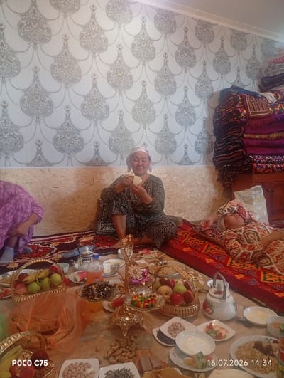 The image depicts an indoor gathering in Чоң-Гара, Kyrgyzstan, with a focus on a spread of food and several individuals. Two women are seated on the floor, one actively eating from her hand, and another lying down with her eyes closed. A third person, partially visible, is seated to the left. The environment features patterned wallpaper and a wooden paneled wall. A table or surface in the foreground is laden with various food items, including fruits in baskets, nuts, sweets, a teapot, and dishes of food, suggesting a meal or celebration. The overall scene conveys a sense of relaxed social gathering.