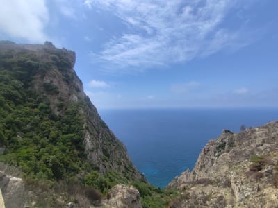 This panoramic image captures a breathtaking coastal landscape from a high vantage point in Λάκωνες, Greece. The scene is dominated by two steep, rugged cliffs that frame a vast expanse of the deep blue sea, extending to a clear horizon.

The cliff on the left is richly covered with dense, lush green vegetation, primarily trees and shrubs, which carpet its slopes from top to bottom. Along the uppermost ridge of this cliff, the weathered stone remnants of what appears to be a historic fortification or castle wall are discernible against the sky, adding a sense of ancient heritage to the natural setting.

In stark contrast, the cliff on the right presents a more desolate and rocky appearance. Its slopes are largely barren, featuring exposed rock formations with only sparse, scrubby vegetation, some of which appears dry or burnt.

Between these two dramatic landforms lies a calm, azure sea, shimmering under the bright daylight. The water's deep blue color transitions to lighter shades closer to the base of the cliffs, suggesting varying depths. The sky above is a brilliant, clear blue, adorned with delicate, wispy white cirrus clouds, indicating fair and sunny weather. The bright illumination suggests a time of day around late morning or early afternoon.

The image evokes a sense of awe and tranquility, highlighting the raw beauty and scale of the Mediterranean landscape. There are no people or discernible text visible in the frame.