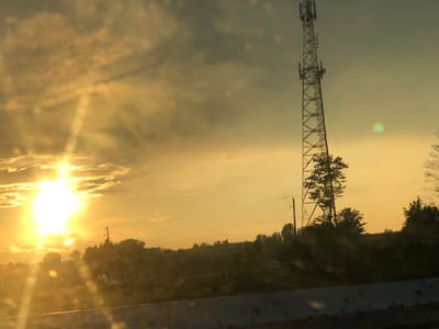 This image captures a rural landscape during a sunset or sunrise. The sun, a bright, glowing orb, is prominent on the left side of the frame, casting a warm, golden light across the scene. Sunbeams cut through the hazy atmosphere, illuminating scattered clouds.
Dominating the right side of the image is a tall, metal transmission tower, characteristic of telecommunication infrastructure. Below the tower and stretching across the middle ground, a line of trees and vegetation forms a natural horizon. Some of these trees are silhouetted against the bright sky, while others are more visible in the fading light. In the distance, gentle hills are visible, hinting at an undulating terrain.
In the foreground, a concrete barrier suggests the image was taken from a road or highway. The lower portion of the image shows a blurred foreground, possibly due to the movement of a vehicle or raindrops on a window, with several circular light refractions scattered across it.
The overall mood is serene and picturesque, with the warm light creating a tranquil atmosphere. The presence of the transmission tower and the concrete barrier juxtaposes the natural landscape with human-made structures. The time of day is clearly indicated by the low angle of the sun and the warm, diffused lighting. There are no visible people in the image. A small, partially obscured green logo with the text "DEERE" is visible on a dark object in the mid-ground on the right side of the image. The location context of Bradford West Gwillimbury, Canada, is not directly evident from the visual elements alone but could provide further context for the type of landscape and infrastructure depicted.