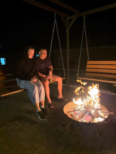 Two people are sitting on a swing next to a fire pit. The fire pit is full of burning wood and the flames are high. The swing is hanging from a metal frame. The ground is covered in dark gray brick. The location is unknown.