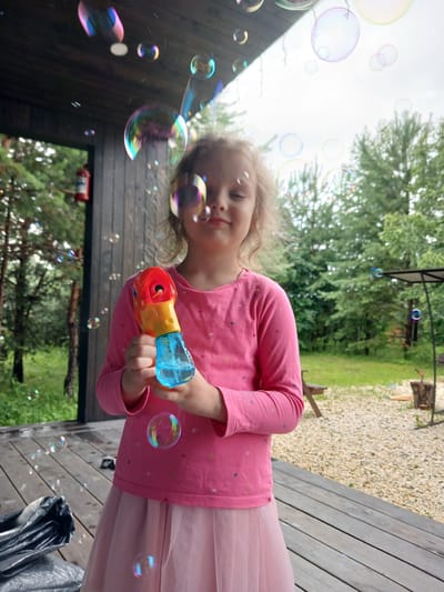 A young girl, appearing to be around 5-7 years old, stands on a wooden porch, holding a red and yellow bubble gun filled with blue liquid. She is wearing a pink long-sleeved shirt with small heart patterns and a pink tulle skirt. She is looking slightly down and to her left, with a gentle smile. The porch is made of wooden planks, and a dark grey bag or tarp is on the floor to the left. In the background, trees and foliage are visible, suggesting an outdoor setting, possibly in a rural or suburban area. The sky is overcast. Numerous soap bubbles of various sizes and colors are floating in the air around the girl and the porch. This scene is taking place in Verkhneye Dubrovo, Russia.