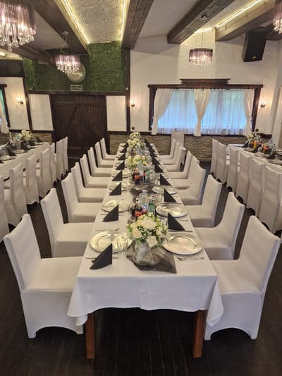 The image shows a long dining table set for an event, likely a wedding, in what appears to be an indoor venue. The location is likely Parvenets, Bulgaria. The table is covered with a white tablecloth, and each place setting features a silver charger plate, a black napkin, silverware, and glassware. Floral centerpieces of white flowers run down the length of the table. White chair covers are on each chair. The room has exposed beams, plaster walls, and a green faux wall with an exit sign above a brown door. A window with sheer white curtains is present. Chandeliers hang from the ceiling. The floors are dark wood. There are no people visible in the frame.