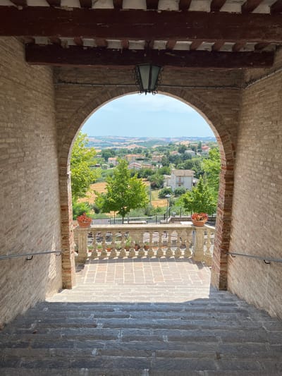 The image shows a view through an arched brick passageway, looking out over a town and surrounding countryside. The passageway is framed by rough-hewn brick walls and a wooden beam ceiling with exposed rafters. A lantern hangs from the ceiling. In the foreground, a stone staircase descends. Beyond the archway, a stone balustrade lines a walkway, with potted plants on top. Trees and buildings are visible in the middle ground, and rolling hills with more scattered buildings make up the background. The sky is clear and blue. The scene appears to be in Barbara, Italy, a small town in the Marche region known for its historical architecture and scenic views.