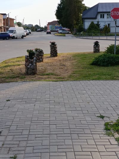 This media file captures a street scene in Smilavicki Selski Savet, Belarus. The foreground is dominated by a gray brick paved walkway, with patches of sparse grass and weeds growing between the bricks. In the middle ground, a well-maintained lawn area features three decorative planters. Each planter is a cylindrical wire cage filled with stones, topped with a dark pot containing small flowering plants and greenery. Beyond the lawn, a paved road curves into the distance, with several vehicles visible. A white van is stopped at an intersection, and other cars are seen in traffic.
On the right side of the frame, a modern white house with a dark roof stands prominently. Next to it, a red octagonal STOP sign is mounted on a pole. The background shows more buildings and trees, suggesting a semi-urban or suburban environment. The lighting indicates it is likely late afternoon or early evening, with soft shadows cast across the scene. The weather appears clear and sunny.
Visible text includes "STOP" on the sign. Additionally, a blue street sign is partially visible in the distance with text that appears to be in a Slavic language, possibly Belarusian or Russian, with the words "ДУКОВА" and "ШАЦК" discernible. No people are clearly visible in the image.