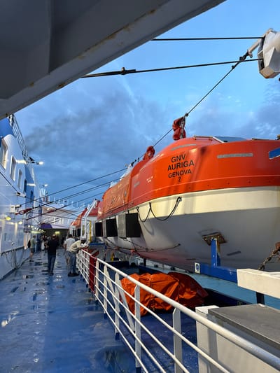The image depicts the deck of a GNV (Grandi Navi Veloci) ferry, specifically the "AURIGA" bound for or docked in Genoa, Italy. A partially visible bright orange and white lifeboat marked "GNV AURIGA GENOVA 2" is attached to the side of the ferry. Several people are walking along the blue-painted deck. White railings are visible along the side of the ship. The sky is partially cloudy, suggesting dawn or dusk. The ferry appears to be docked next to another vessel. It is presumed the ferry is in Civitavecchia, Italy.