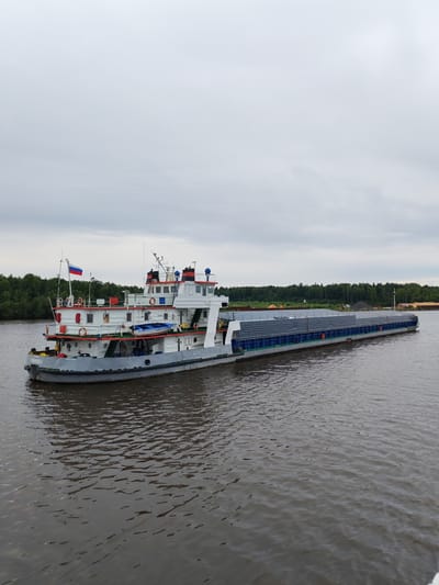 The image shows a barge being pushed by a tugboat on the Desyatovskaya canal in Russia. The barge is loaded with what appears to be steel pipes. The tugboat has a Russian flag on its mast. The canal is surrounded by trees on both sides. The sky is cloudy. The water is dark and choppy.