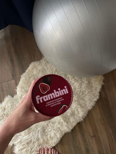 A photograph taken from an overhead perspective presents a scene indoors in Sepych, Russia. A person's hand holds a cylindrical container, the lid of which is predominantly maroon in color and labeled with a white inscription that reads "Frambini." The lid also features an image of a dark chocolate-covered raspberry.
Adjacent to the hand and the container is a plush, light-colored rug. In the background, a silver-colored exercise ball is partially visible. The floor is made of wooden planks, and the individual's toes are observable on the right side of the photograph. The person is wearing navy blue pants.