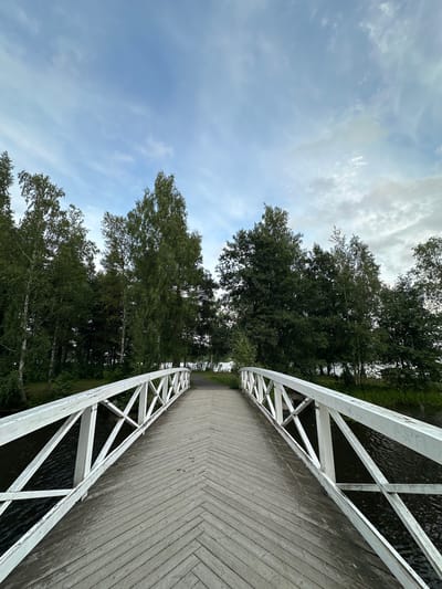 The image depicts a wooden bridge with white railings, leading over a body of water. The bridge deck is constructed of light-colored wooden planks laid in a herringbone pattern. The railings are white and feature a latticed design with diagonal supports. Trees line the banks on either side of the water, with lush green foliage. The sky above is partly cloudy, with patches of blue and wispy white clouds. The overall scene suggests a peaceful, natural setting, likely a park or waterfront area. The location is identified as Vammala, Finland. There are no people visible in the image, and no discernible actions or interactions are occurring.