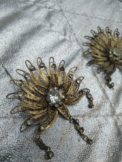 The scene is located in the city of Тохта, Kazakhstan. The image presents a close-up, static view of two ornate, flower-shaped metal ornaments, likely brooches or hair adornments, resting on a silvery, textured fabric surface.
The primary subject, located prominently in the foreground, is a roughly 10-12 cm wide ornament crafted from tarnished, possibly brass or bronze-toned metal, featuring intricate filigree-style petals. This piece exhibits significant oxidation and wear. A large, clear, round faceted rhinestone forms the center, surrounded by numerous smaller clear, round faceted rhinestones embedded within the metallic petals. Three separate chains with small, dark bead-like terminals dangle from the lower edge of this ornament.
A second, similar but smaller and less distinct ornament is partially visible in the upper right background. It also appears tarnished, with embedded clear stones, and is less in focus than the foreground item.
The background consists of a reflective, silvery-grey fabric, possibly satin or a similar material, which displays subtle wrinkles and folds, suggesting a soft, uneven surface. Minor dark particulate matter is visible on the fabric. No individuals, active interactions, or further environmental details are present in the image.