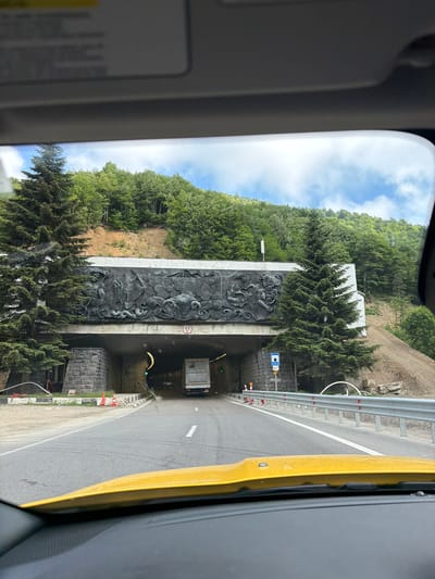 The image shows a view from inside a vehicle, looking out at a road leading to a tunnel entrance. The tunnel entrance is large and arched, with stone masonry on the sides. Above the tunnel entrance is a large, dark bas-relief sculpture depicting figures and possibly animals, set against a white background. There are pine trees on either side of the tunnel entrance, with a dense forest covering the hillside behind it. A truck is visible entering the tunnel. The road has white lane markings and is bordered by a metal guardrail on the right. In the foreground, the yellow dashboard of the vehicle is visible. The sky above is blue with some clouds. This appears to be the entrance to the Surami Tunnel in Surami, Georgia.