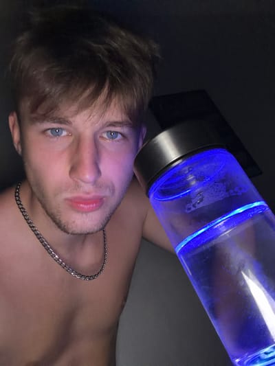 The image depicts a close-up, waist-up shot of a young adult male with fair skin, blue eyes, and light brown hair. He is shirtless and wearing a silver chain necklace. He is holding a clear glass bottle with a silver cap, filled with a blue-lit liquid, in his right hand, bringing it close to his face. The background is dark and indistinct. There is no information in the image to suggest a location in the city of Vanino, Russia.