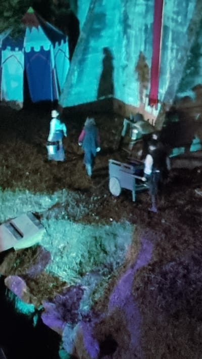 The image depicts an outdoor scene, possibly at a historical reenactment or festival, given the presence of tents and figures in what appear to be period attire. In the background, two large, tent-like structures are visible, one predominantly blue with white accents and the other a lighter color with a red vertical stripe. The lighting is atmospheric, with a prominent teal and purple hue cast over the scene, suggesting artificial lighting.

In the mid-ground, three individuals are present. Closest to the viewer and moving away is a person wearing a blue garment and what appears to be a white head covering. To their right and slightly further back, another individual is seen from the back, wearing a dark vest over a lighter colored garment and dark pants. To the right of this second person, a third individual, dressed in dark clothing and a white top, is operating a small, two-wheeled cart.

The ground is covered in what looks like dry grass and soil, with some areas illuminated by the teal and purple light, creating an ethereal effect. A flat, rectangular object, possibly a piece of equipment or signage, lies on the ground in the lower left quadrant of the image. The overall impression is one of a staged event taking place in an outdoor setting, possibly at the historic mills of Sant Joan les Fonts, Spain, given the suggestion of a rural or heritage location.