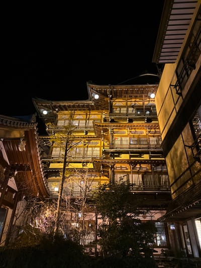 The image depicts a multi-story, traditional Japanese building, illuminated at night, situated in Yamanouchi, Japan. The structure features prominent tiered roofs with distinctive upward-curving eaves, adorned with multiple circular light fixtures emitting warm yellow light. Each floor is characterized by numerous windows, predominantly covered by light-colored curtains, and is fronted by an extensive wooden balcony or walkway with intricate railings. The building's exterior is predominantly a muted yellow, complemented by dark wooden structural elements.

To the left, a portion of another traditional building with a curved roofline and dark wooden siding is partially visible. To the right, a cream-colored building with dark horizontal trim lines and a contrasting lighter roof overhang frames the scene. In the foreground and midground, a dense cluster of deciduous trees and shrubs, some with sparse foliage, are present. These plants are irregularly illuminated, casting shadows and highlighting individual branches. The sky above is uniformly dark, indicating nighttime. No people or active subjects are visible within the frame.