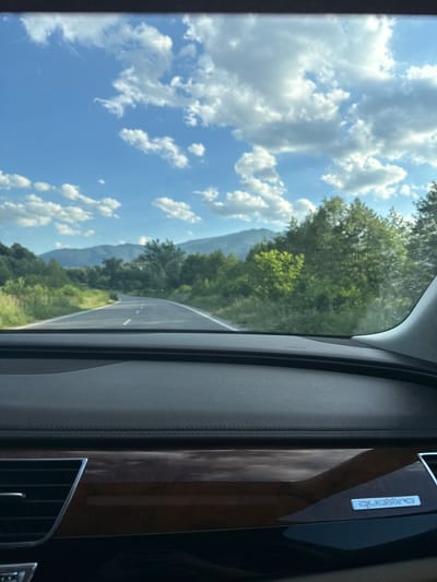 The image, taken from inside a car, shows a view through the side window. Outside, there is a blue sky with fluffy white clouds. A winding road is visible, surrounded by trees and mountains. The car's interior is visible, including the panel next to the window. The wood panel has a silver "Quattro" badge on it. The location is potentially near the city of Mezdreya, Bulgaria.