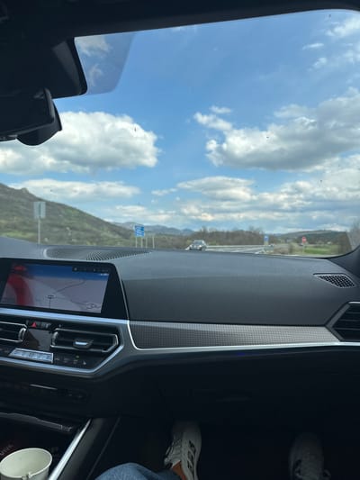The image shows the interior of a car, likely a BMW, with a view out of the front windshield. The dashboard and infotainment screen are visible, displaying a navigation map. Through the windshield, a road stretches into the distance with a mountainous landscape and a partly cloudy sky. There is no evidence of Bregovo in the image. There is a person visible with their feet and part of the lower body. A white cup is in the lower right corner.