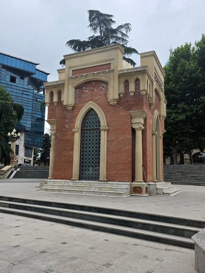 The image shows a small, ornate brick structure with a decorative arched doorway and stained-glass windows, situated in an open, paved public area in the city of Ganja, Azerbaijan. The structure appears to be a mausoleum or a historical monument. It is surrounded by stone steps leading up to its entrance, and a large, modern building with a glass facade is visible in the background to the left. Trees and a green dome with a statue on top are visible in the background to the right, suggesting a park or a religious site nearby. The sky is overcast, and the overall lighting is subdued. There are no people visible in the immediate vicinity of the structure. The foreground is a paved plaza with distinct square paving stones and darker granite steps.