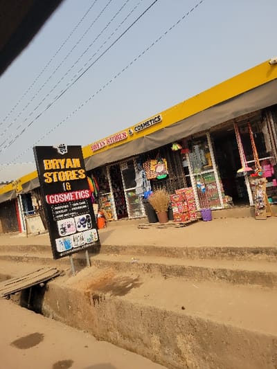 The image depicts a commercial street scene in Puerto López, Colombia, during daylight. A single-story building features a yellow-painted facade with an illuminated sign displaying "BRYAN STORES & COSMETICS". A black, freestanding sign on the unpaved ground to the left reads "BRYAN STORES 16 COSMETICS", listing items such as perfumes, skin care, jewelry, and promotions, with corresponding product images. The storefront consists of multiple glass display windows showcasing various retail items, including brooms with colorful handles, bags, and stacked boxes of merchandise. Two dark planters containing dried vegetation and a purple bucket are positioned on the ground outside the store. A concrete open-air gutter runs parallel to the building in the foreground, bridged by a wooden plank in the lower left. Beyond the gutter, a raised concrete walkway or series of steps leads to the store entrances. Numerous thin electrical wires crisscross against a clear, light blue sky overhead. No individuals are visible in the frame.
