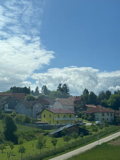 The image shows a view of a residential neighborhood in Kirchstetten, Austria, at coordinates 48.187 latitude and 15.823 longitude. The scene is captured from a high vantage point, likely from a vehicle. The sky is bright blue with large white cumulus clouds. The foreground is a grassy slope with a paved road curving through it. There are several houses, mostly two-story structures, with a variety of colors, including yellow, grey, and red. The houses are arranged in a loose cluster, with a few trees and shrubs interspersed between them. The trees are mostly deciduous, with a few evergreens visible in the background. The houses are in good condition, with well-maintained lawns and gardens.  A white car can be seen parked on the road.