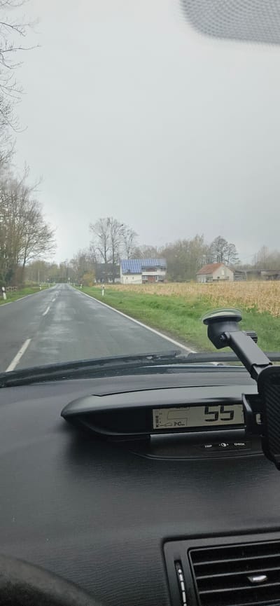 The image, taken from inside a vehicle, shows a view of a wet road leading towards the distance. In the background, on the left, there is a house with solar panels on its side. Further in the distance, there is a field and another building. The speedometer in the vehicle reads 55. The photo was taken in Rietberg, Germany.