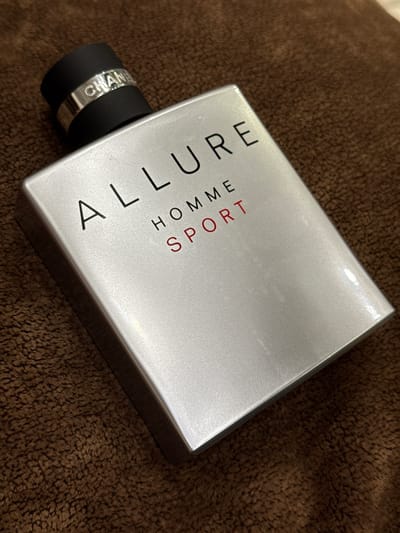 The image shows a silver perfume bottle lying on a brown, textured surface, possibly a towel or carpet. The bottle is a rectangular prism with rounded edges and a black cap. Text is visible on the bottle. The largest text, oriented diagonally, reads "ALLURE SPORT". Below this, in smaller, vertical text, it says "HOMME". Around the black cap, there is a silver band with "CHANEL" inscribed on it. The lighting suggests an indoor setting, with some reflections on the bottle's surface indicating a light source. No people are present. The overall scene is a close-up, still life of a luxury perfume product.