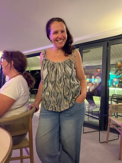 The image shows two women indoors, likely in a restaurant or bar in Grimaud, France. One woman is standing, smiling directly at the camera, wearing a patterned tank top and jeans. The other woman is seated, leaning forward towards a table, wearing a white t-shirt. The standing woman is holding the back of the seated woman's chair. In the background, there's a window or glass door reflecting an outdoor scene with greenery and what appears to be other patrons. The environment is brightly lit with a mix of natural and artificial light. There's a table with plates and chairs visible.
