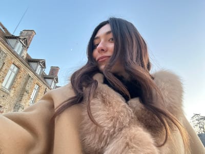 A female individual is captured in a low-angle selfie, looking upwards and slightly to the right, with long dark brown hair framing her face. She is wearing a light beige coat with a prominent light-colored fur or faux-fur collar over a dark garment. The background features a historic stone building characteristic of the Abbaye des Vaux de Cernay in Cernay-la-Ville, France. The building displays multiple windows, dormer windows on a pitched roof, and chimneys, constructed from light-colored stone. A clear, light blue sky dominates the upper frame, featuring a visible crescent moon. To the far right, a line of trees is silhouetted against the horizon.