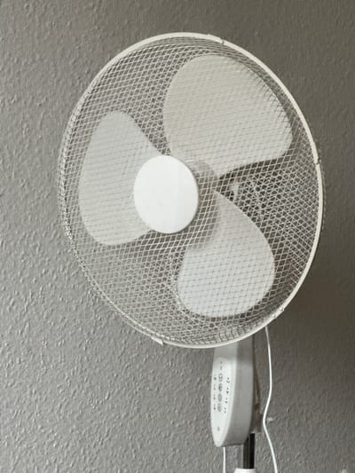 The media file displays a white electric fan with a circular mesh grill, positioned in a close-up view against a textured gray background. The fan has three white blades, and the central hub is also white. On the right side of the fan, a white control panel with several buttons and indicator lights is visible. Some of the buttons have small icons, suggesting different fan speeds or modes. A white power cord extends from the control panel, disappearing out of frame. The lighting appears to be artificial, likely from an indoor setting, and there are no clear indicators of time of day or weather. The scene is static, with the fan appearing to be either off or not in operation, as there is no motion blur. The textured background suggests a wall or ceiling. The overall impression is a functional household appliance captured in a simple, straightforward shot. No people are visible in the frame. The location context provided is Bad Blankenburg, Germany, but this cannot be visually confirmed from the image itself.