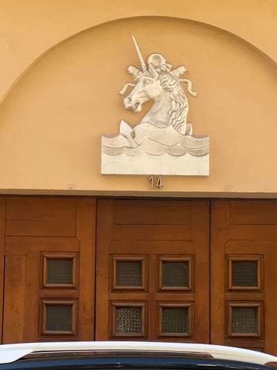 A stucco relief of a unicorn emerges from stylized waves above a pair of wooden doors. The unicorn is white, with a spiraling horn, a mane, and flowing ribbons. Below the relief, the number "14" is displayed in silver metal numerals. The wooden doors are paneled, with six square glass inserts on each door. The glass inserts appear to be leaded or patterned. The doors are set within an arched alcove, and the wall above the alcove is painted a pale orange. A white car is partially visible in the foreground, below the doors. The architectural style suggests a building in Toulon, France, possibly dating from the mid-20th century. There are no people visible in the image.
