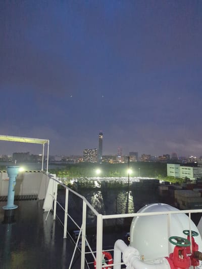 The image shows a view from the deck of a ship at night, looking towards the city of Chiba, Japan. The sky is dark and cloudy, with a few faint lights visible in the distance, possibly stars or distant aircraft. In the foreground, there is a railing and part of a white dome-shaped object with red valves and pipes. The deck is wet, suggesting recent rain.

In the middle ground, there is a line of trees separating the ship from a brightly lit area that appears to be a parking lot filled with cars. Beyond the trees and the parking lot, the city skyline is visible, with numerous buildings illuminated by artificial lights. A tall, slender skyscraper with a red light at its peak stands out prominently. The city lights create a hazy glow in the night sky.

The overall scene depicts a port or industrial area in Chiba, Japan, during nighttime, likely shortly after a rain shower. The focus is on the transition from the industrial setting of the port to the urban landscape of the city. There are no people visible in the image.