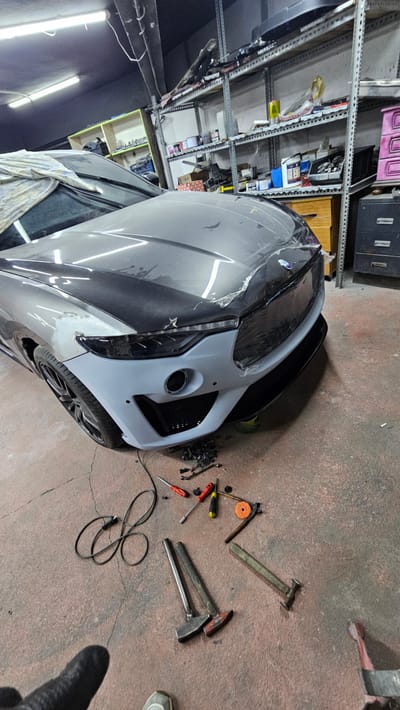 The image captures an indoor workshop setting, likely a car repair or customization shop, located in Ataşehir, Turkey. In the foreground, a grey luxury SUV is partially covered with protective plastic sheeting and a cloth, with its front bumper removed and replaced with a primer-coated replacement part. Various tools, including hammers, screwdrivers, and a polishing pad, are scattered on the concrete floor around the vehicle, suggesting ongoing repair work. In the background, metal shelving units are filled with car parts and supplies, typical of a professional automotive workshop. The overall impression is one of active work in a cluttered but organized industrial space.