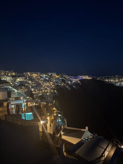 This is a nighttime, high-angle view of a cliffside town with white buildings and numerous lights. The town is situated on a hillside that slopes down towards a distant city illuminated by many lights. The architecture is consistent with Cycladic style, characterized by whitewashed buildings with cubic forms and flat roofs. Some buildings have balconies and terraces, and many windows are illuminated, suggesting occupancy or active businesses.

In the foreground, there are outdoor seating areas with tables and chairs, some of which are illuminated by warm-toned lights. A prominent structure features a neon sign that reads "ROCCA" in white illuminated letters, with "NOAH" below it in smaller, white illuminated letters. Adjacent to this, a turquoise-lit pool or water feature is visible, surrounded by a woven bamboo-like fence. Further in the foreground, there are more roof structures and architectural elements, some with wooden railings and beams. The sky is dark blue, with a faint glow on the horizon from the distant city. The overall impression is of a vibrant nightlife scene in a beautiful coastal town. Based on the architecture and general appearance, this scene is likely in the town of Fira, on the island of Santorini, within the Thira Municipal Unit, Greece.