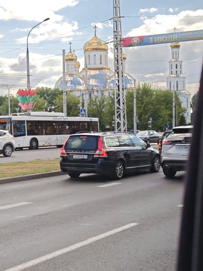 In Alexandrovka, Russia, a black Volvo V70 wagon is parked on a street, its rear facing the camera. The car's license plate is visible and reads "2140 HA-1". To its right is a white Ford Edge SUV, also parked. In the background, a white trolleybus with the text "Любимый Брест" (Beloved Brest) written on its side is stopped, with passengers visible inside. Further in the background, several domed buildings, likely churches, stand tall against a cloudy sky. A banner with Russian text, including "3 ИЮЛЯ" (July 3rd) and "ДЕНЬ НЕЗАВИСИМОСТИ РЕСПУБЛИКИ" (Independence Day of the Republic), is stretched across the sky between two poles. Trees and traffic signals are also present in the scene.