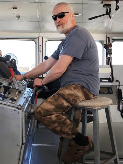 A gray-haired man with a beard and sunglasses sits on a stool in what appears to be the cockpit of a ship or boat. He wears a gray t-shirt and camouflage pants, with brown shoes visible on his feet. He is positioned at the controls, with his hands on levers and switches, suggesting he is operating the vessel. The cockpit is predominantly gray, with various control panels and equipment visible. Through the windows, a view of land and water is discernible. The scene appears to be indoors.