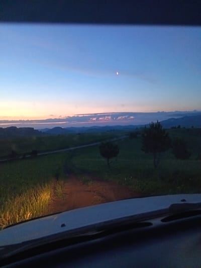 The image depicts a rural landscape at dusk, viewed from inside a vehicle. The sky is a gradient of deep blue to pale pink near the horizon, with a faint crescent moon visible. Rolling hills and distant mountains form the background, shrouded in twilight. In the mid-ground, a dirt road winds through a grassy field dotted with trees. The foreground is dominated by the interior of the car, specifically the dashboard and windshield, with the headlights illuminating the tall grass at the edge of the road. The overall impression is one of peaceful solitude in a natural setting. The location is identified as Sadovyi, Russia, implying a rural or semi-rural environment within that region.